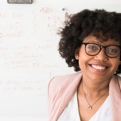 Close up photo of a smiling woman facing the camera with a whiteboard full of writing behind her.