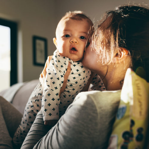Photo taken over the shoulder of a mother sitting sideways on a couch, kissing her baby on the cheek as they look at the camera.