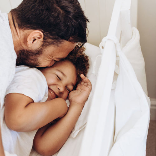 Dad and young son, both wearing white t-shirts, snuggle in a bed with white furniture and white sheets.