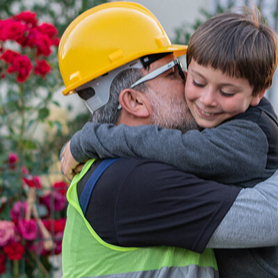 Working man hugs his son before leaving for work. Kneeling in front of their family home and garden.